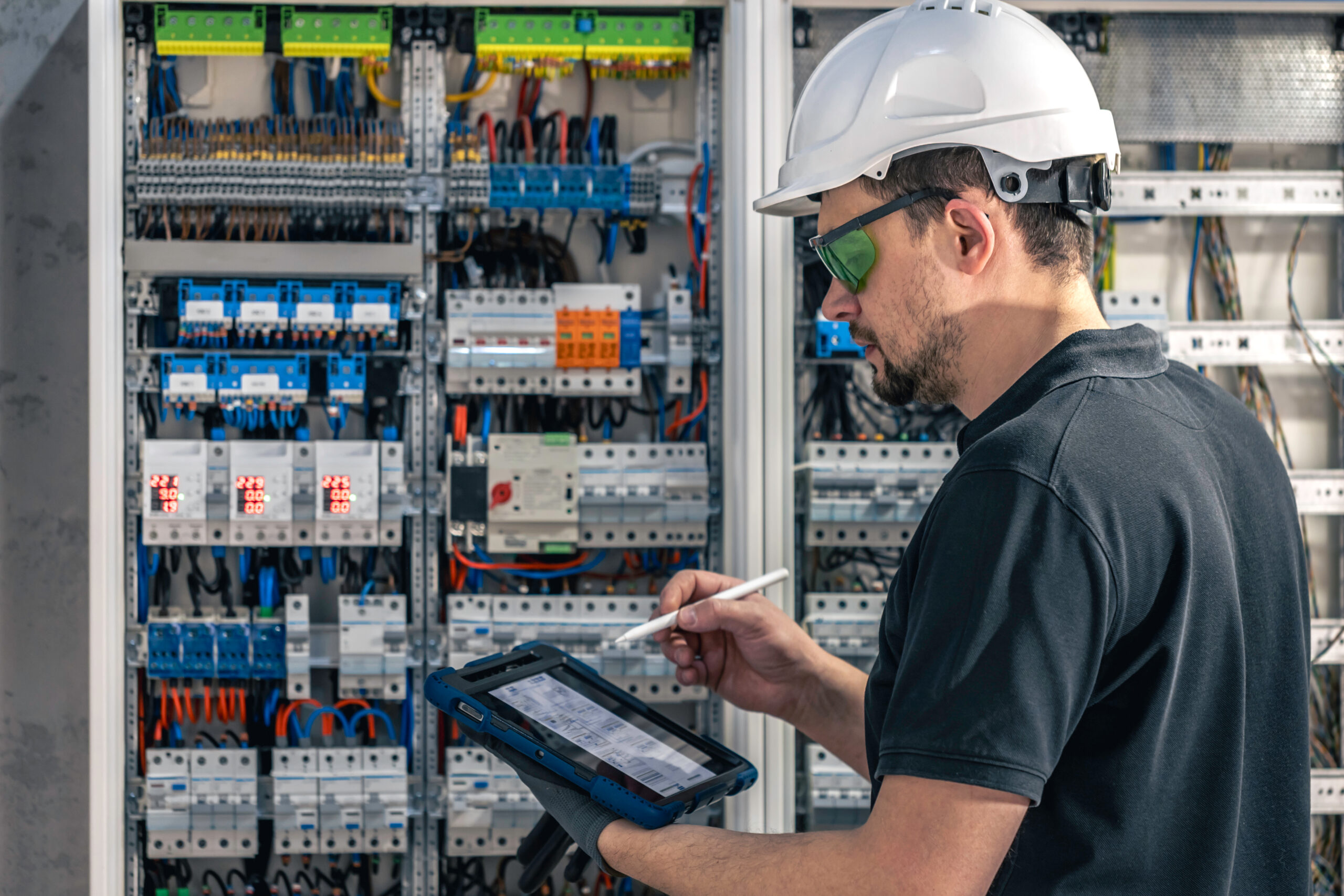 Man, an electrical working in a switchboard with fuses, uses a tablet.